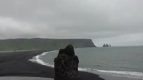Waves rolling in over black sandy beach with steep cliffs and rocky pillar a Stock Footage 136892426