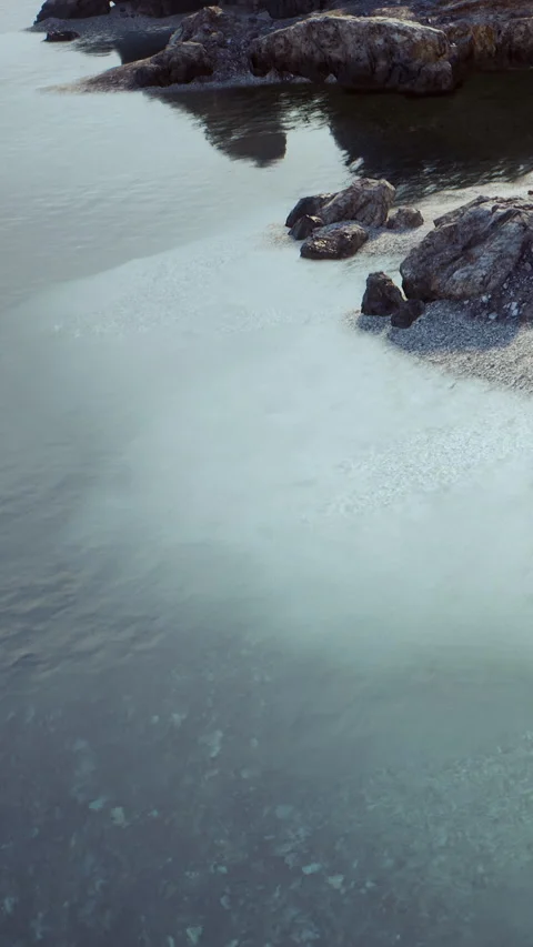 Waves touch rocks on the shore while water reflects light in clear skies Stock Footage 340639001