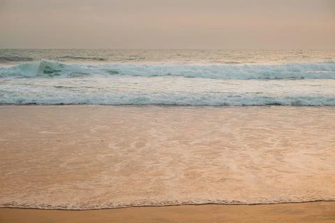 Waves wash ashore on sandy beach during evening light near ocean. Waves roll Stock Photos