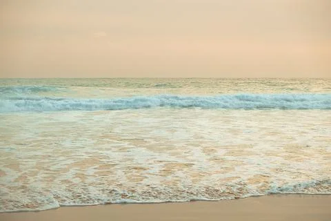 Waves wash ashore on sandy beach during evening light near ocean. Waves roll Stock Photos