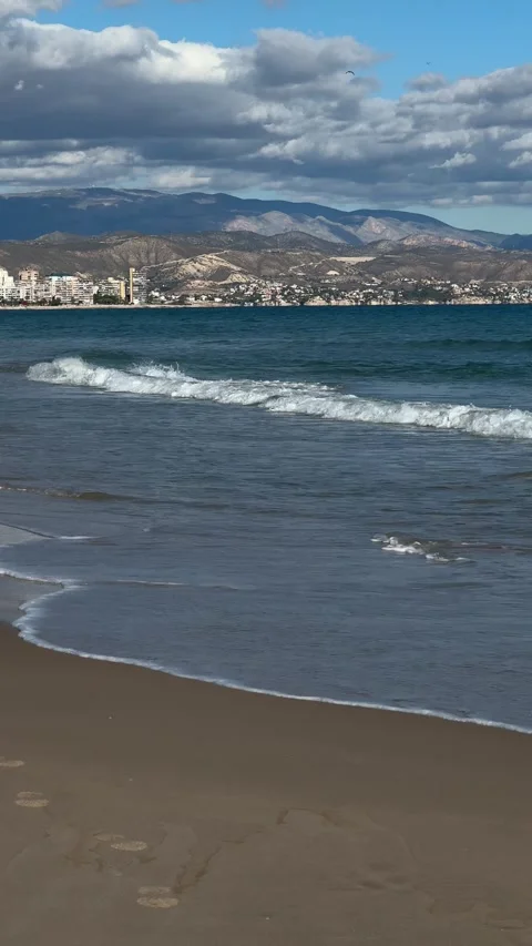 Waves washing sandy shore with distant mountains and urban coastline in Alicante Stock Footage 295205658