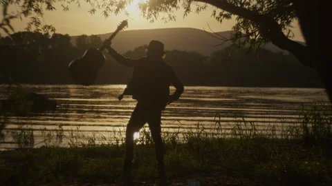 Waving man plays guitar by the river at sunset in peaceful setting Stock Footage 319952479