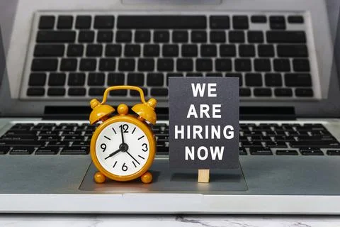 We are hiring now text on chalkboard with clock set at 8 o'clock on a laptop. Stock Photos