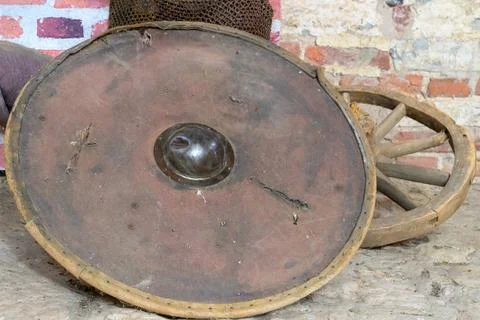 Weathered circular shield rests beside wooden cartwheel. Rusty metal center Stock Photos
