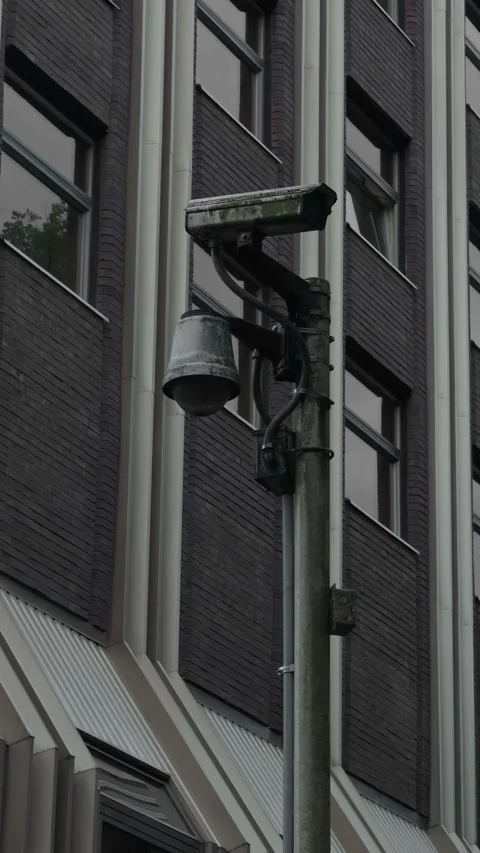 Weathered security camera mounted on modern building facade, angled toward urban Stock Footage 337647913