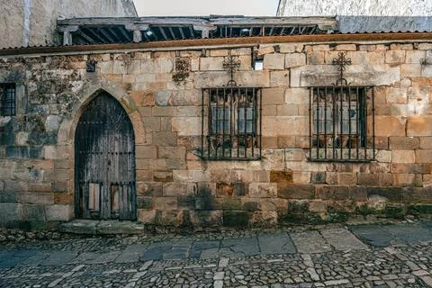 Weathered stone facade featuring an old arched wooden door and two barred win Foto stock