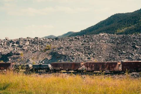 A weathered train sits before a vast rock pile and verdant mountain side. Stock Photos