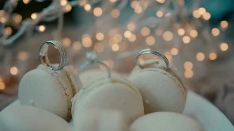Wedding rings and an engagement ring stuck in macaroons with lit garlands Stock Footage 221781939