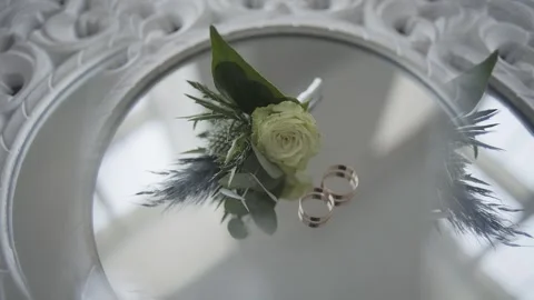 Wedding rings lie on a mirror next to a natural flower boutonniere on the Vídeos de archivo 306145019