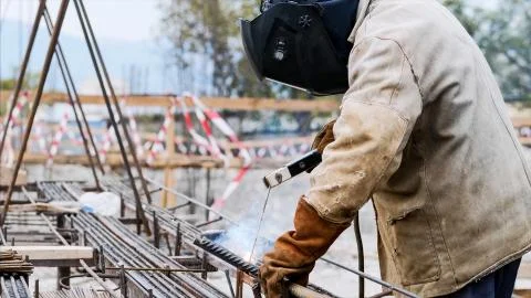 Welder at construction site is welding pipe with electrode in helmet. Stock Photos