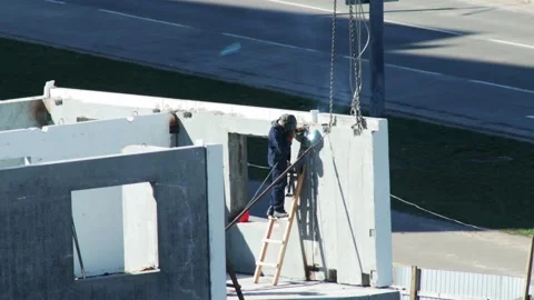 Welder On Ladder Secures Concrete Panel Joint. Second Worker Climbs Ladder To Stock Footage 323912935