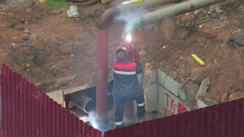Welder in protective uniform performs repair work on large heating main pipes Stock Footage 344864225