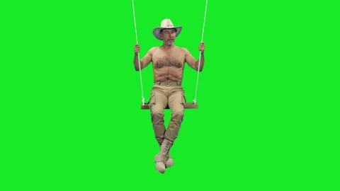 A well-built man in cowboy hat and boots swinging on rope swing on green screen Video stock 305190068