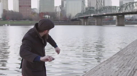 Well-dressed man skipping rocks on a river off of a dock in the city by a bridge Stock Footage 60071968