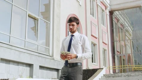 A well-dressed man stands outside a contemporary building, counting cash with a Stock Footage 316043747