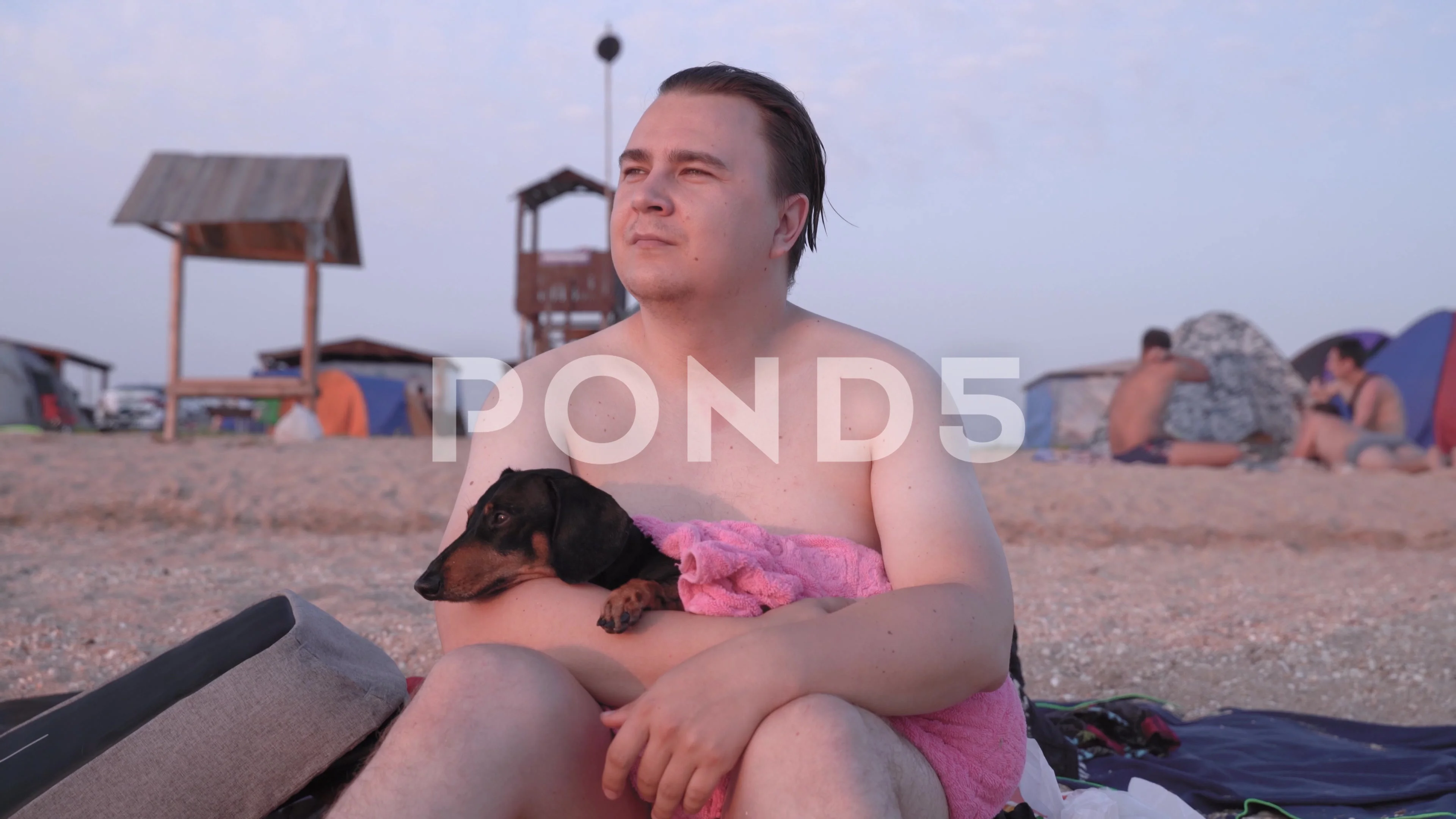 Wet naked guy sits with dog on his laps on sandy beach after swimming.