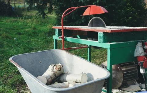 A wheelbarrow filled with cut logs placed beside a manual sawing machine in.. Stock Photos