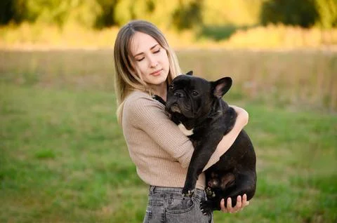 While walking in park, young dog owner holds her beloved pet, French bulldog, in Stock Photos
