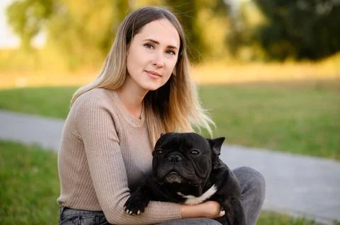 While walking on sunny summer day, happy woman sits and cuddles her black French Stock Photos