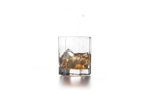 Whiskey splash from the fallen ice cube into glass with beverage on light bac Stock Photos