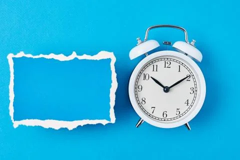 White alarm clock and empty torn paper sheet on a blue background, top view.  Stock Photos