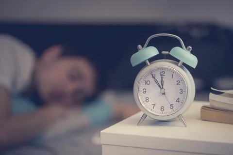 White alarm clock on the bedside table. Young man sleeps in the background Stock Photos