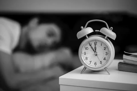 White alarm clock on the bedside table. Young man sleeps in the background Stock Photos