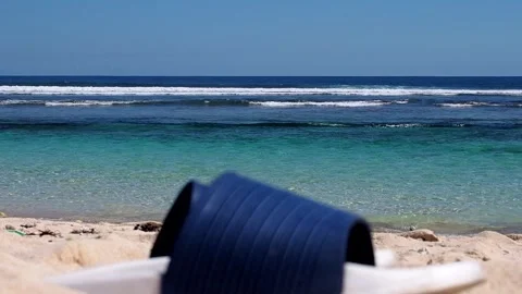 White and blue flip flops on a white sandy beach against a blue ocean backdrop Stock Footage 248530737