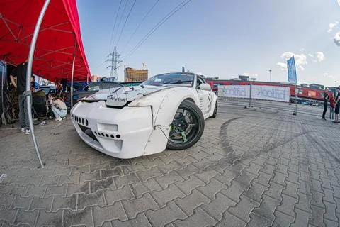 White angular BMW car with front-end damage on pebbled ground, near a red c.. Foto stock