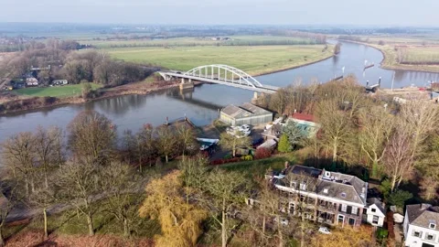 A white arched bridge spans a calm river curving through open fields Stock Footage 328680443