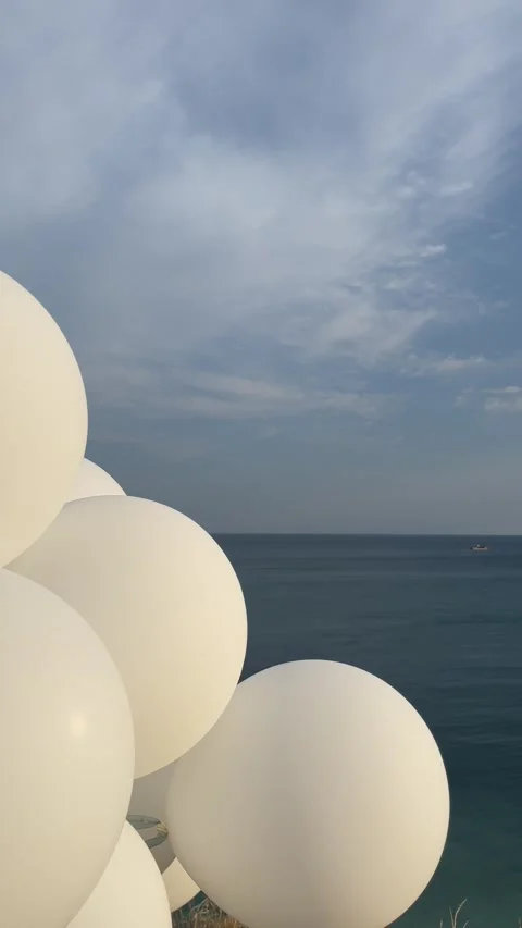 White balloons flutter in the wind against a serene sea a blue cloudy sky Stock Footage 283066875