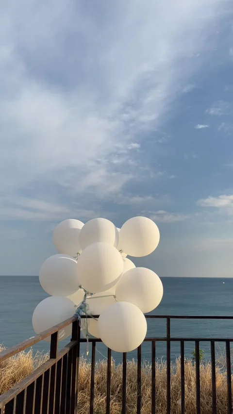 White balloons flutter in the wind against of a serene sea and a blue cloudy sky Stock Footage 283066889