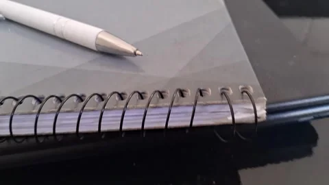 White ballpoint pen resting on grey spiral notebook video footage Stock Footage 339739846