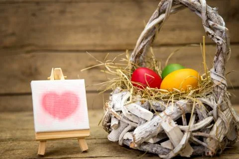 White basket with easter eggs, canvas with red heart, wooden background 库存照片