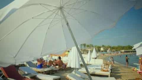 White beach parasol providing shade over a beach lounger, with people relaxing, Video stock 343204269