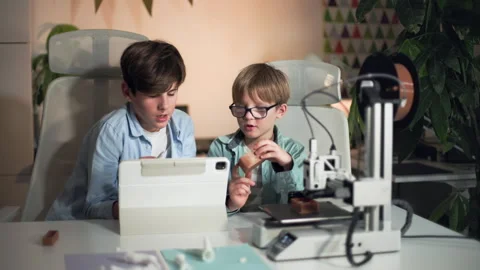 At a white bright table, two kids control 3D printer and discuss printed part Stock Footage 335378607