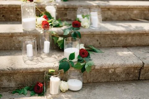 White candles in glass jars surrounded by roses stand on stone steps Stock Photos