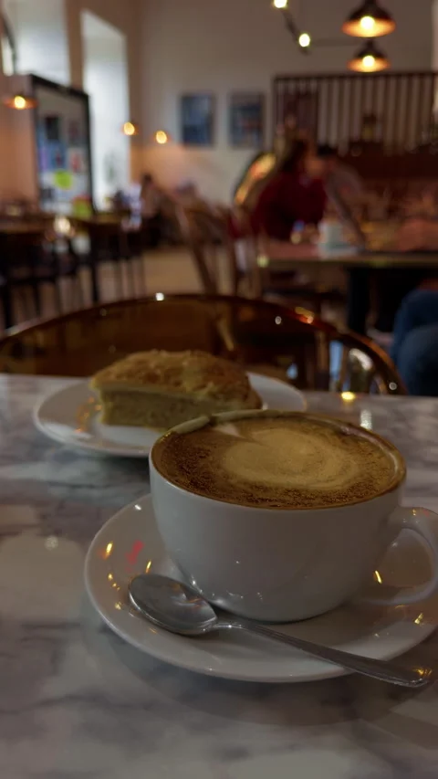 White cappuccino cup with spoon beside slice of cake on marble cafe table in Stock Footage 344966893