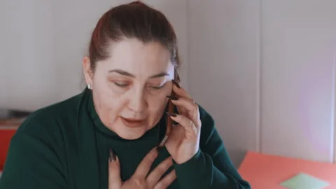 White caregiver holding chest while pleading on phone in home setting, tearful Video stock 342336853
