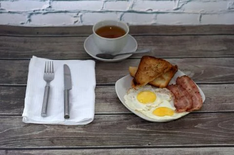 A white ceramic plate with two fried eggs, toast and bacon is on the table, n Stock Photos
