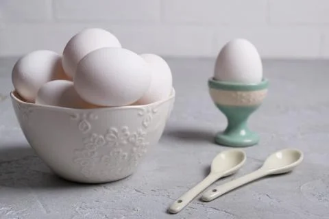 White  chicken  egg in ceramic stand and eggs in white  bowl . On a gray Foto stock