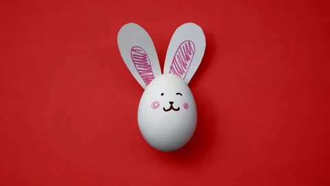 White chicken egg with cute bunny face, paper rabbit ears on light red Stock Footage 302522110