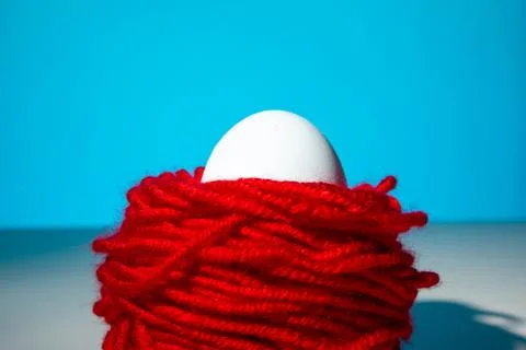 White chicken egg in red threads blue background, close up, copy space Stock Photos