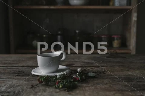 A White China Cup And Saucer On A Kitchen Table And A Twig Of Pine And Berrie