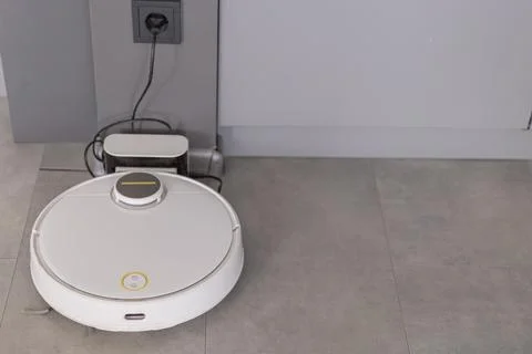 White circular robot vacuum cleaner charging on gray tile floor near Stock Photos