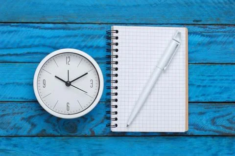 White clock and notebook on blue wooden background. Minimalistic studio shot. 库存照片