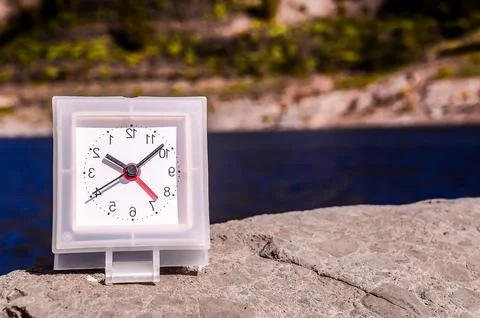 A white clock with red hands sits on a rock by a body of water Foto stock