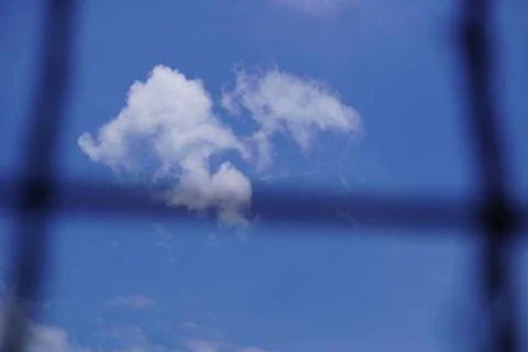 White cloud in blue sky viewed through a blurred dark metal wire mesh fence f Stock Photos
