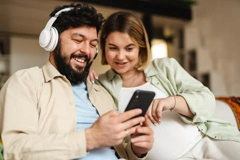 White couple using cellphone and headphones while sitting on couch Foto stock
