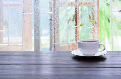 A white cup of coffee is placed on a table by the window. Stock Photos